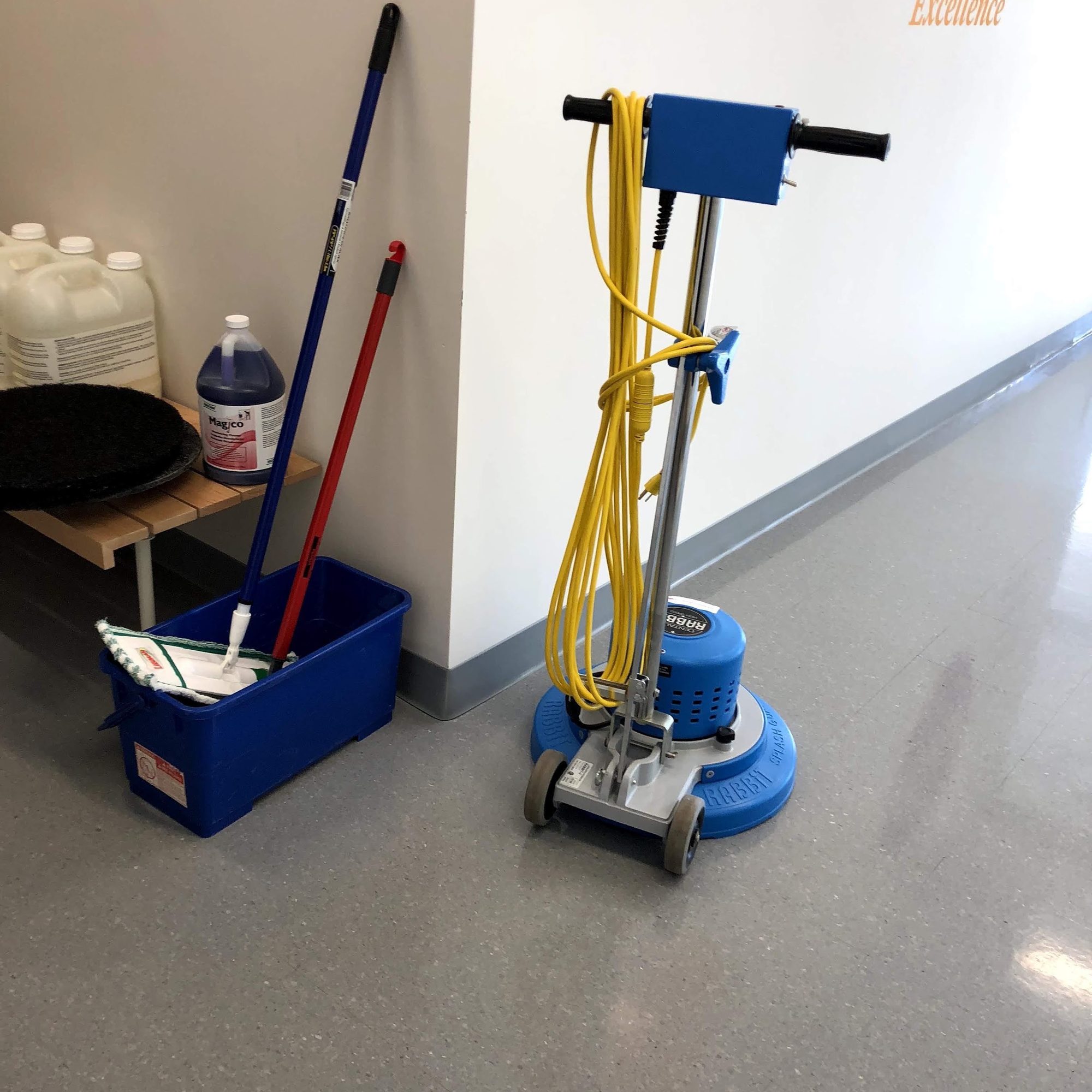 A rotary floor machine, mop bucket and cleaning products lined up against a wall