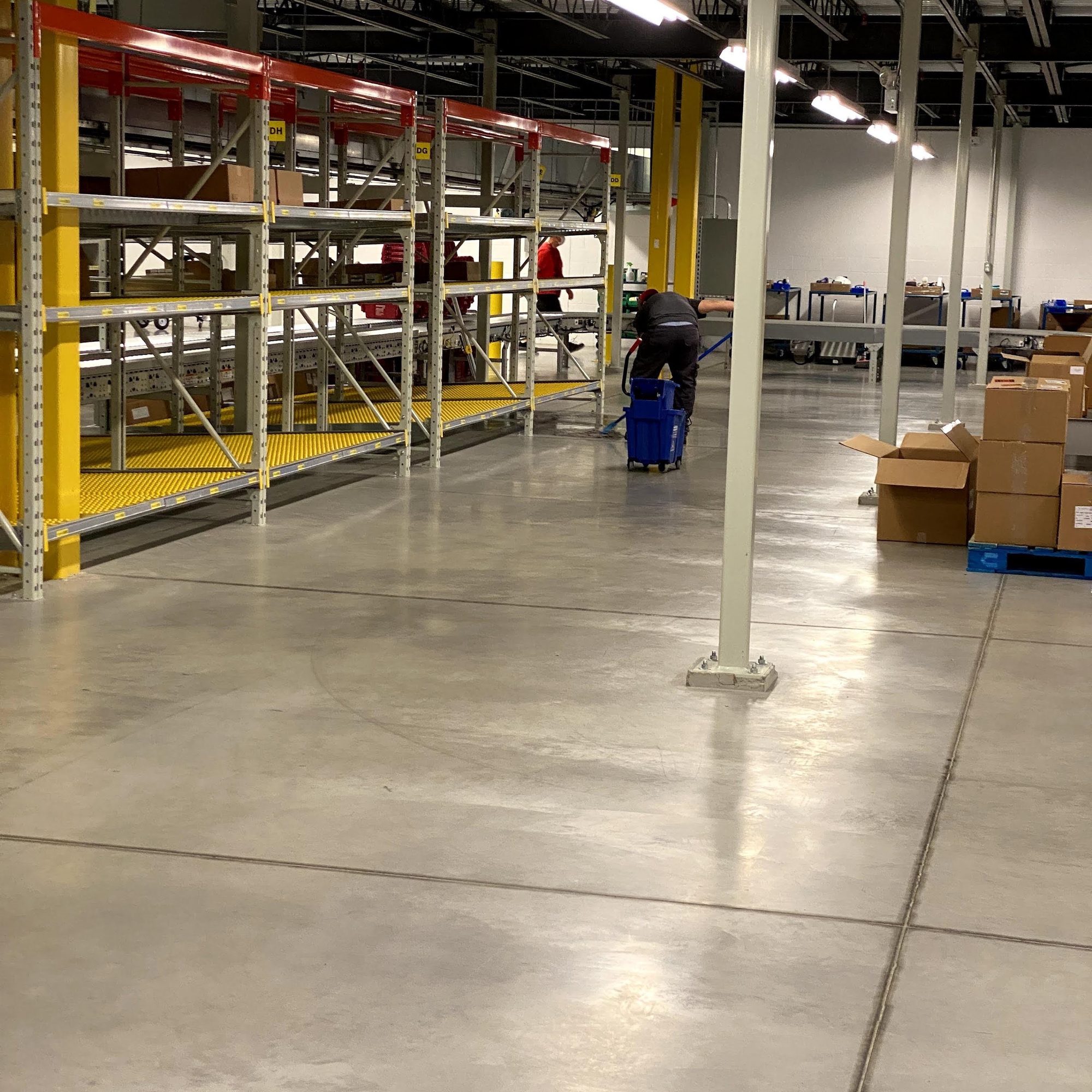 Polished concrete floor in a distribution centre beside yellow racking, a cleaner mopping in the background